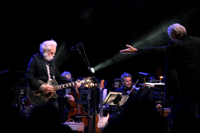 A man with long white hair and mustache plays an intricate black guitar. Beside him, a conductor gestures to the symphony.