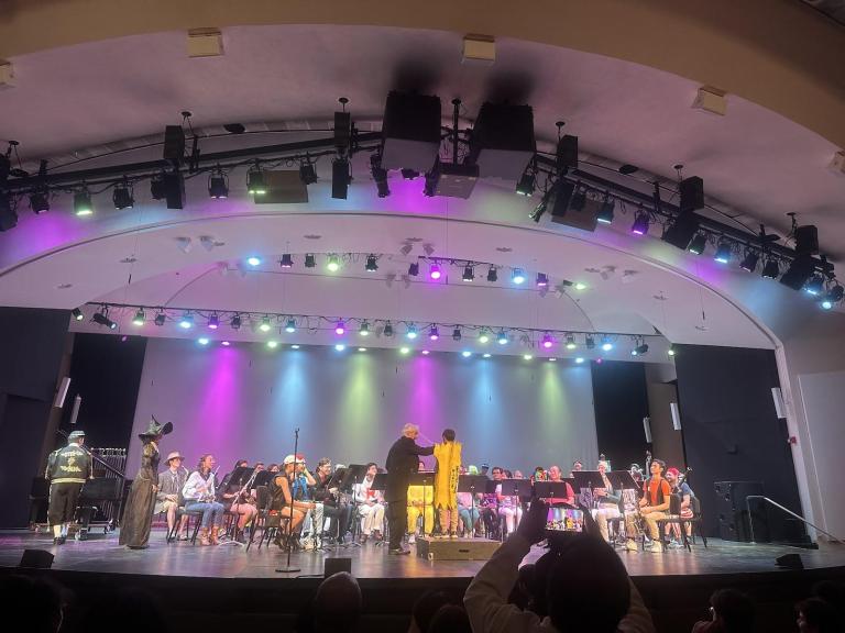 a photo of a stage decked out in pink, blue and yellow lights, with musicians and a person in a boxy yellow costume