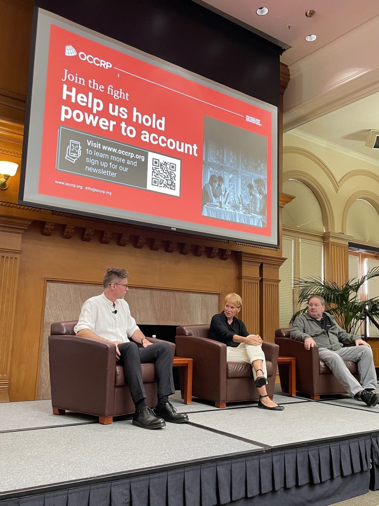 Three speakers sit in chairs under a large screen presenting slides