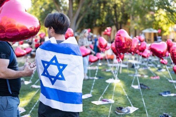 A boy stands with an Israeli flag wrapped around his back. In front of him are dozens of heart shaped balloons attached to pairs of shoes.