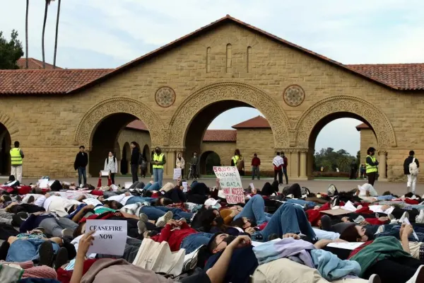 About 30 students, some with masks and some with signs, lie down on main quad. Observers and safety officers in yellow vests stand nearby.