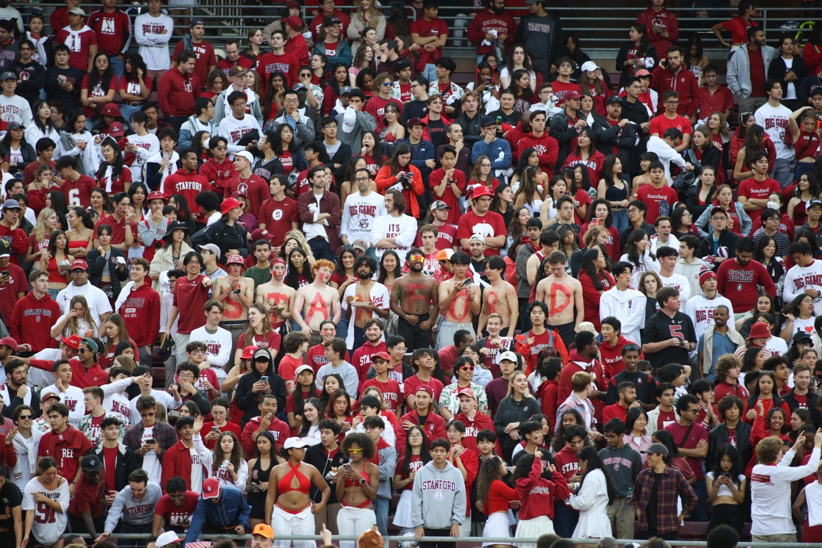 Stanford fans in the stands. Eight students stand shirtless with "STANFORD" spelled in red paint across their chests.