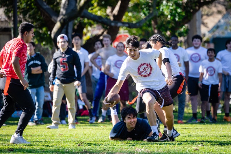 A player in a Stanford Daily shirt leaps over a Daily Cal player as others watch from the sidelines.