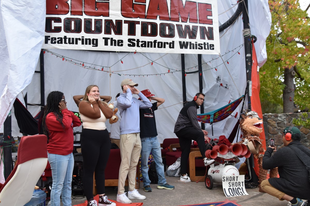 Axe committee members cover their ears under a sign reading "Big Game Countdown Featuring the Stanford Whistle." One wears noise-cancelling ear muffs and prepares to honk the horn while another films.