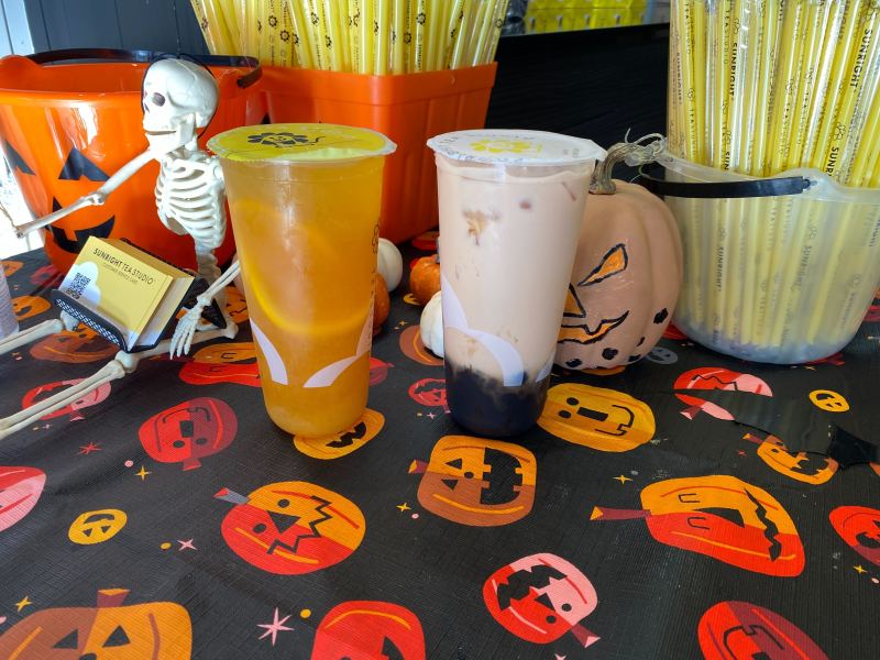 Two drinks, one light orange and the other off-white with brown boba at the bottom. They sit on a halloween tablecloth in front of a pumpkin and a skeleton.
