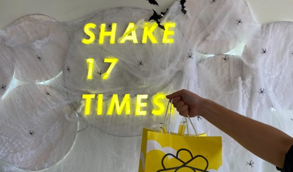 A yellow shopping bag held in front of a white wall decorated with spiderwebs. On the wall is a neon sign reading "Shake 17 Times."