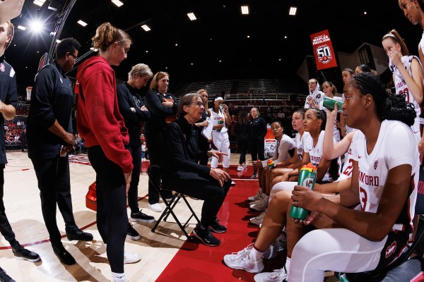 Tara Vanderveer sits in a chair courtside talking to players seated and standing around her.