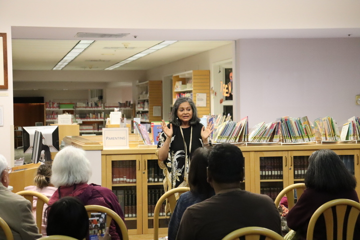 Storyteller Roopa Mohan talks to an audience in a library with shelves stocked full of books behind her.