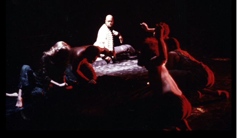 College-aged Andre Braugher onstage in the spotlight. Other students crawl toward him, lit in red.