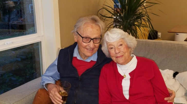 Rowland and Patricia Rebele on a couch. They're leaning into each other and Rowland has one arm wrapped around Patricia and a cup in another.