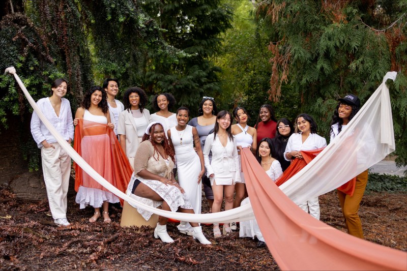 Several non-white students wearing white and pastel colors stand next to each other in front of a wooded background.