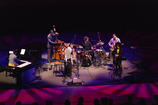 Terri Lyne Carrington singing into a microphone at Bing Concert Hall with her ensemble, all illuminated with warm lights of blue and pink.