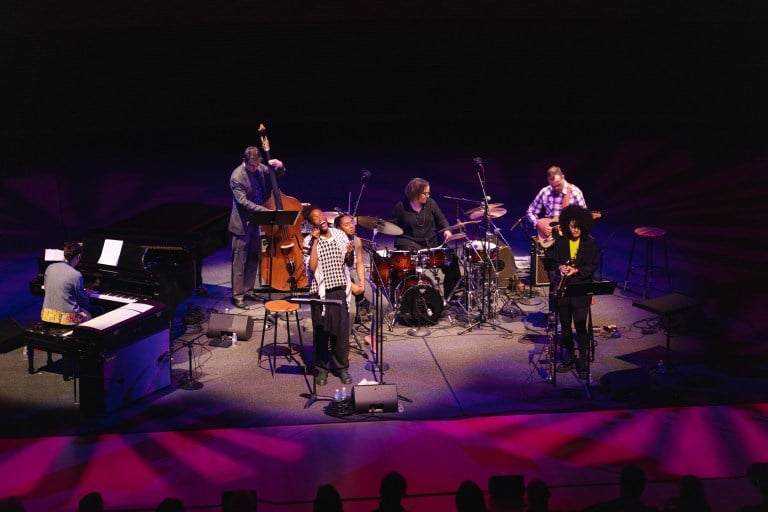 Terri Lyne Carrington singing into a microphone at Bing Concert Hall with her ensemble, all illuminated with warm lights of blue and pink.