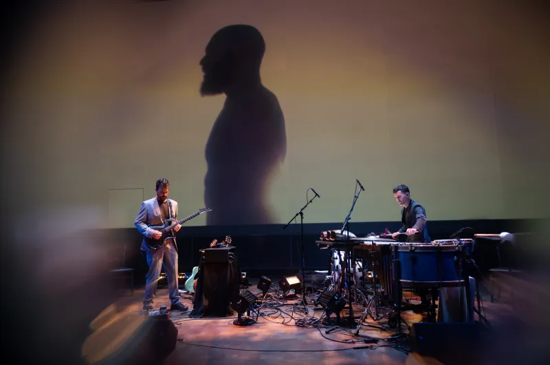 Two musicians playing on stage. A man's shadow is projected onto the wall.