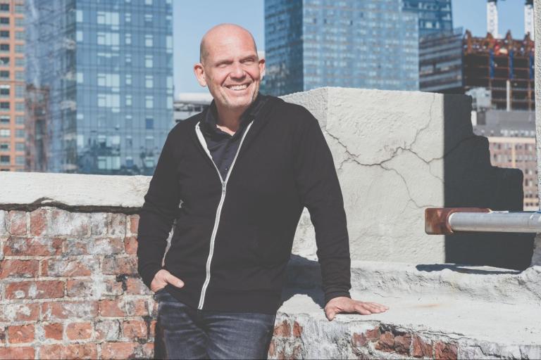 Jaap van Zweden, clad in black jacket and blue jeans, leans against brick wall on building roof.