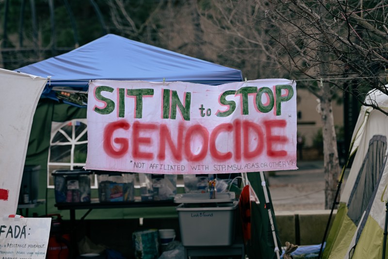A banner hanging on a tent in White Plaza reads, "Sit In to Stop Genocide."