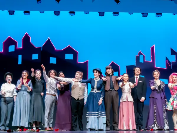 Photo of a cast on a stage, in a performance of Mary Poppins. Most are wearing dresses.