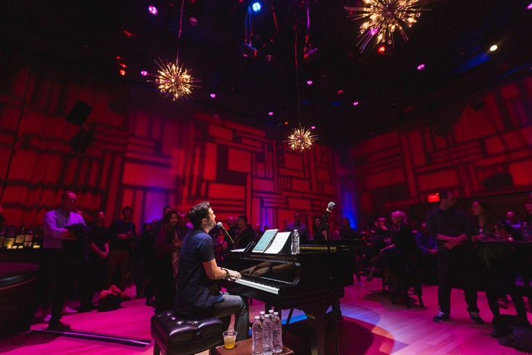 A white man plays a piano in a small venue as people stand around him. The room is dim and lit with red lights.