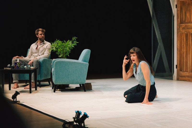 A white woman sits on the floor, distressed, as a white man sits in a chair looking at her. The set resembles a modern therapist's office