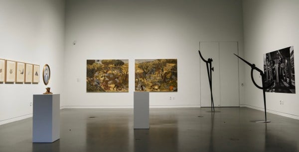 A small art gallery featuring two sharp, large metal sculptures and two small metal sculptures. On the walls are two large colorful paintings, a black and white photograph, and some small paintings.