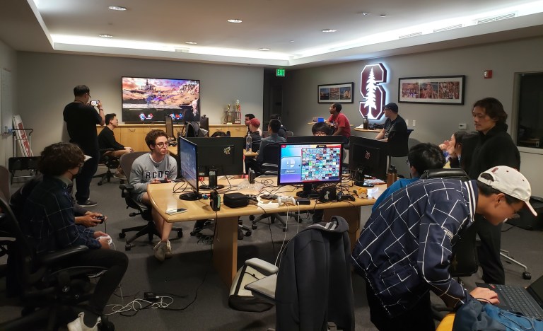 A table with monitors, displaying Super Smash Bros. title. and members of the group playing titular game