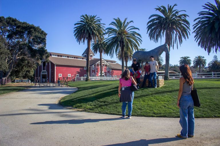 A family of four in front of a white horse statue in front of the Red Barn and two people walking towards the frame.