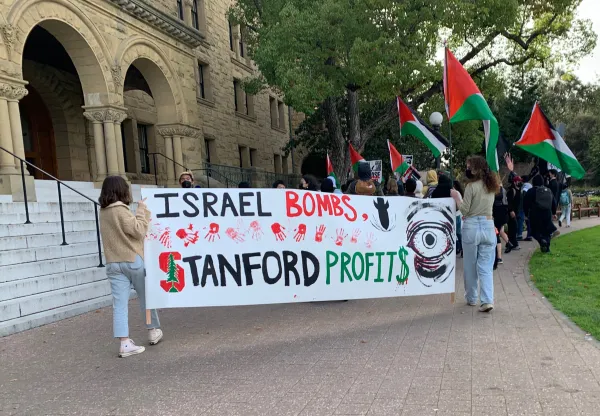 Students in front of Encina Hall carrying a banner that reads 'Israel bombs, Stanford profits.'