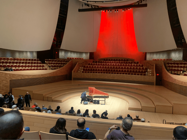 A harpsichord sits at the center of the Bing Concert Hall stage with a bright spotlight shining upon it, red lights in the background.