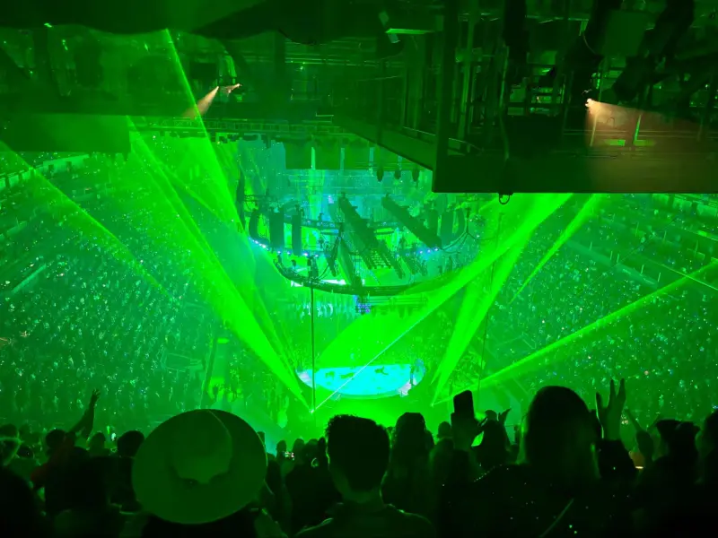 Green lights shine up from a hemisphere-shaped stage in Chase Center of San Francisco, illuminating silhouettes of audience members waving their arms to the music.