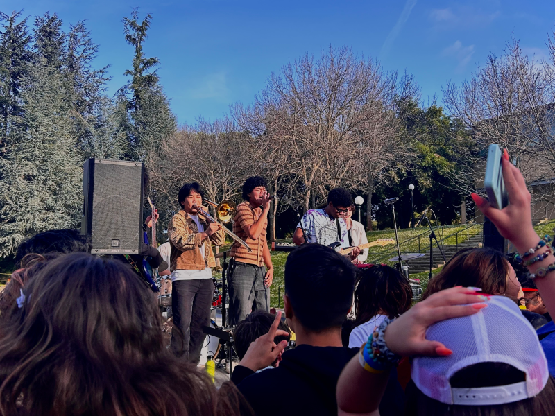 Six of Spades band member perform on a stage in front of Terman Fountain and a crowd of students.
