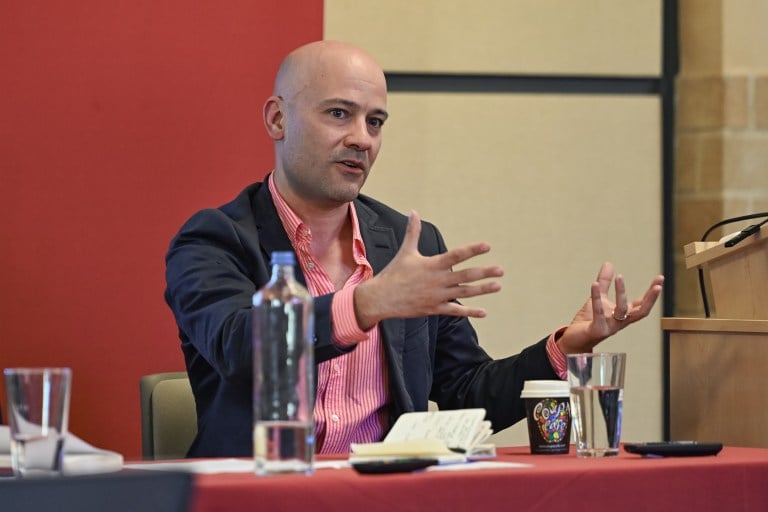 A man addresses an audience from a seat at a conference room table.