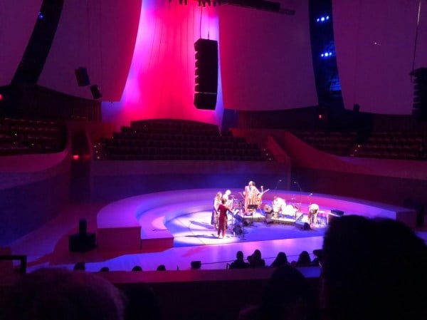 A group of musicians stand in a semi-circle on the stage of Bing Concert Hall with musical instruments from a variety of different cultures. The stage is lit with bright pink and violet lighting.