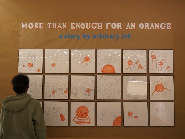 A student stands in front of a brown wall at On Call Cafe. The wall displays a series of 20 panels with pen-and-ink illustrations of a brightly-colored anthropomorphic orange, drawn in a storybook style. The words "More than enough for an orange: a story by Arbitrary Ink" is displayed above the drawings.