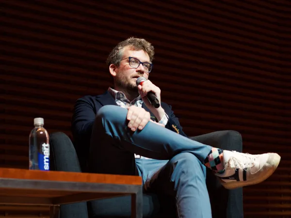 John Green sits comfortably, holding a microphone, as he addresses an audience.