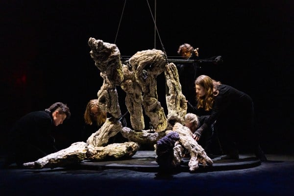 A large puppet, which appears to be the figure of a giant made out of wood, is sprawled across a dark stage. Five puppeteers dressed in black surround the puppet and are partially hidden from view.