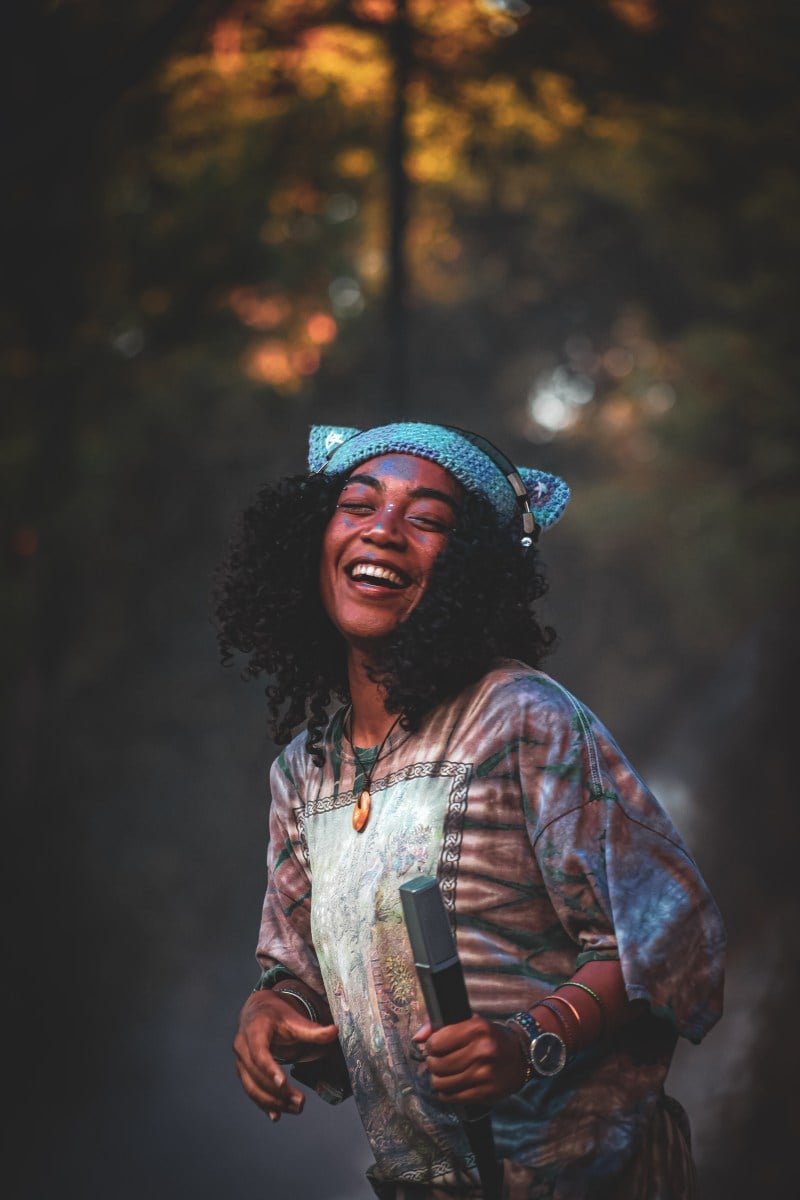 A performer smiles at the crowd. She wears a blue hat and over the ear headphones. Her face is smeared with flecks of blue paint.