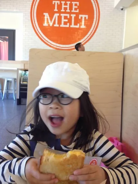 A young Dan Kubota eating grilled cheese in a white cap with orange "The Melt" logo on the wall behind her