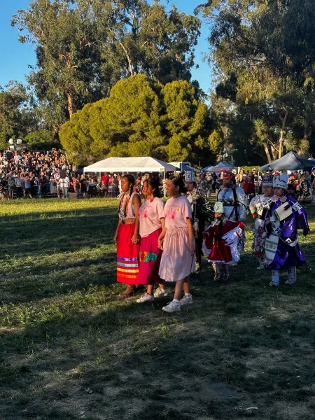Three female students wearing bright pink "Powwow" T-shirts walk hand-in-hand in a grass field. Women dressed in Native American traditional dress, called regalia, walk behind them.