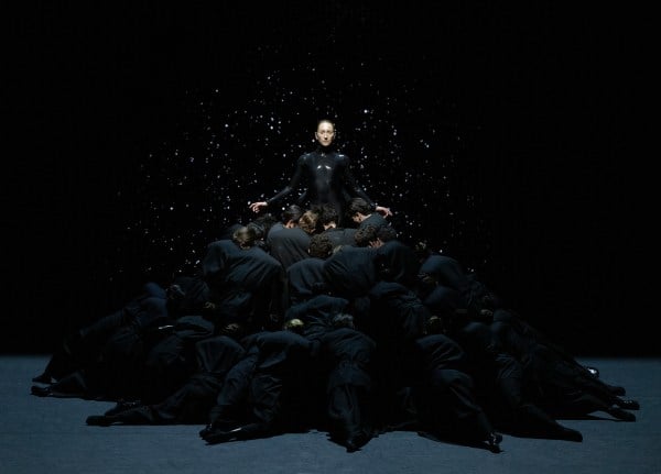 A woman in a shiny black bodysuit calmly rises above a pile of people in black jumpsuits. In performance of Mere Mortals.