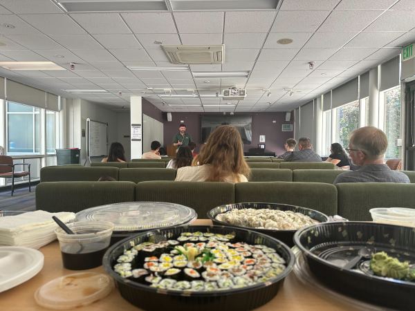 Plates of sushi are on a table in the lecture room of SLE filled with sofas of students and a lecturer talking to them.