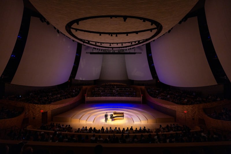 Liu stands next to the piano in a full concert hall.