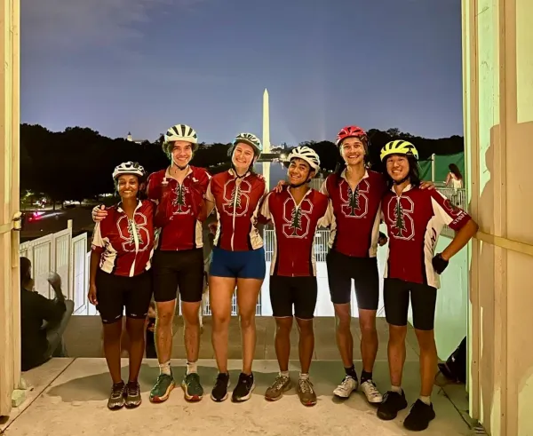 All six members of the 2024 Stanford Spokes Team stand in front of the Washington Monument at night, wearing bike helmets and matching red Stanford shirts.