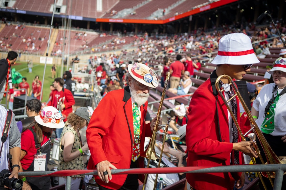 Members of the Stanford marching band climb stairs with the Stanford football field in the background.