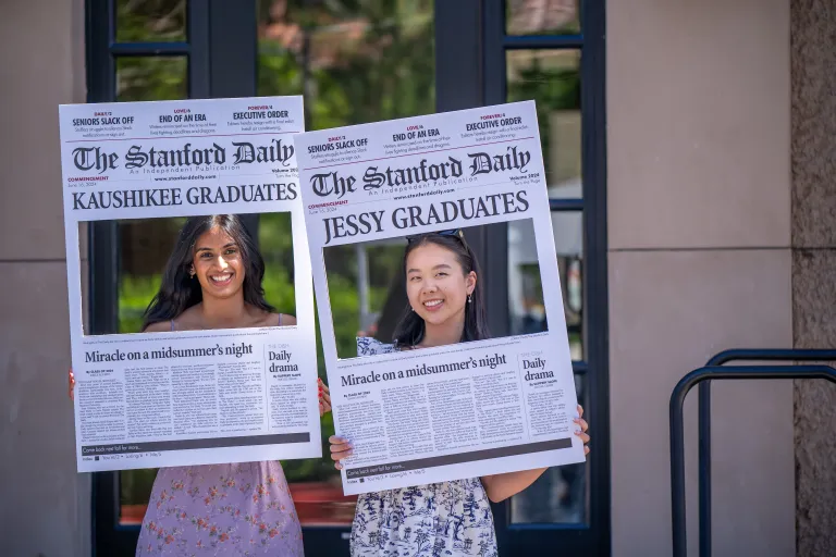 Two individuals hold up cardboard cutouts that mimic a newspaper but with headlines that reads Kaushikee graduates and Jessy graduates.