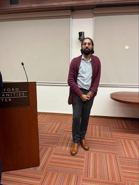 Mohr Visiting Poet L. Lamar Wilson poses in Levinthal Hall, donning a maroon cardigan on a light grey button-up, black pants and brown dress shoes.