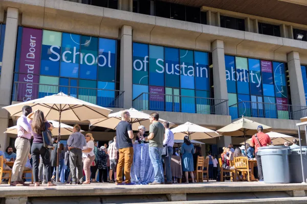 People congregate around tables outside of the Doerr School. A banner on the building in the background reads "School of Sustainability."