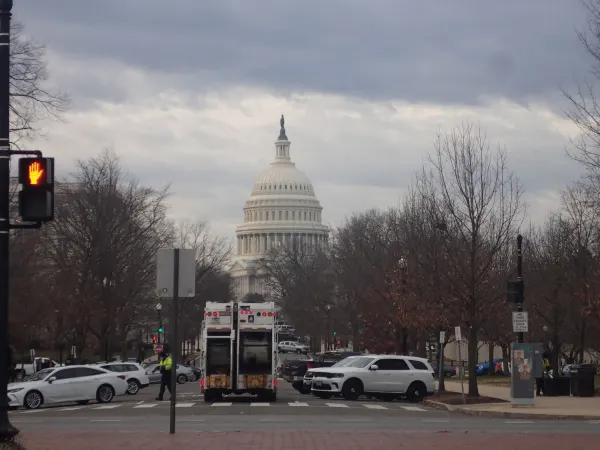 The Capitol rises under a cloudy sky. Cars are parked along the street.