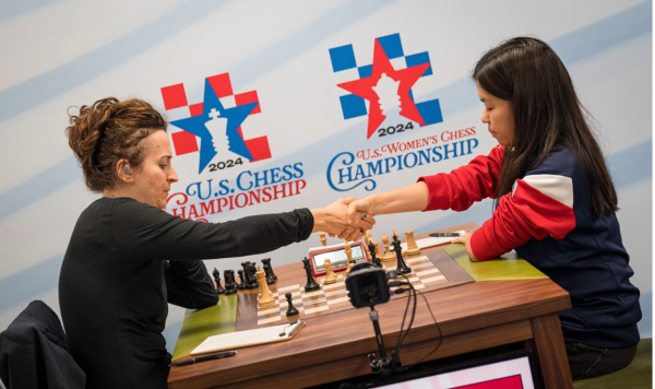 Two women sitting across from each other and shaking their hands over a chess board.