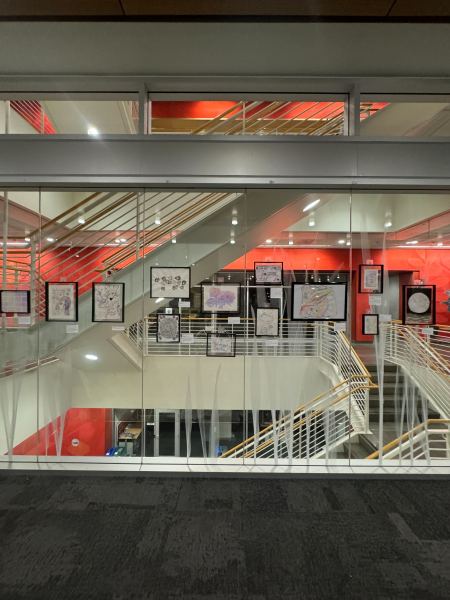 Photo taken of the biopigment paintings along a glass transparent wall with a stairwell visible on the other side.