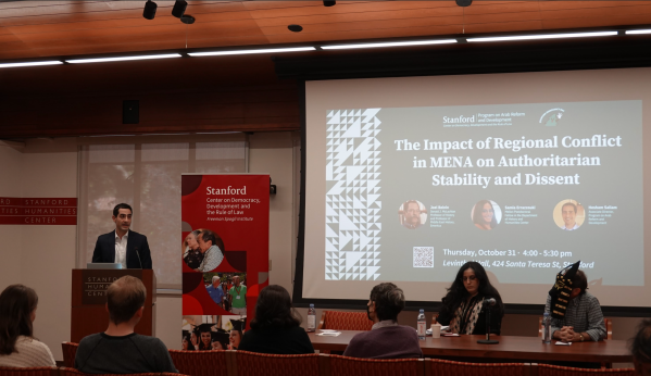 A speaker stands at a podium in front of several audience members, while two other speakers sit at a table, one of them wearing a Halloween mask. A slideshow behind the speakers reads, "The Impact of Regional Conflict in MENA on Authoritarian Stability and Dissent."